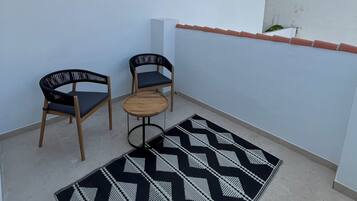 Un pequeño balcón con dos sillas de madera y una mesa redonda pequeña, un tapete estampado y una pared blanca.
