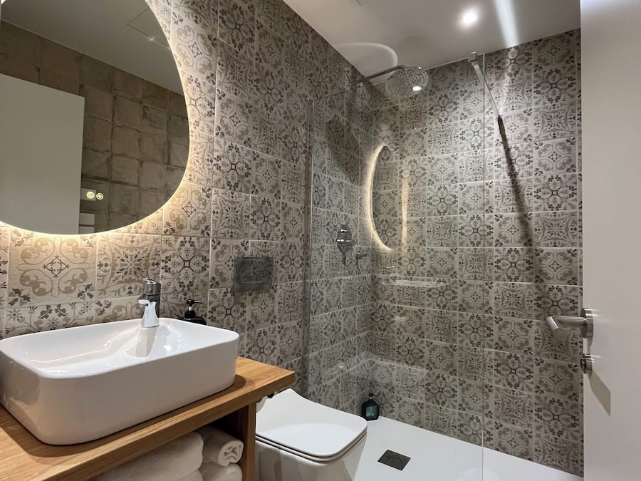 A modern bathroom with a large oval mirror, a white sink, a toilet, and a glass-enclosed shower.