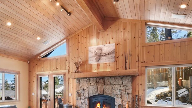 A spacious room with a stone fireplace, wooden ceiling, large windows, and a view of the outdoors.