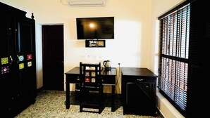 A small, compact room with a TV mounted on the wall, a desk with a chair, a small cabinet, and a window with blinds. - Napier Heritage (Kochi)