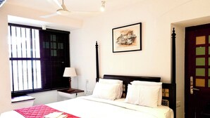 A hotel room with a bed, a window with blinds, a bedside table with a lamp, and a door with a decorative panel. - Napier Heritage (Kochi)