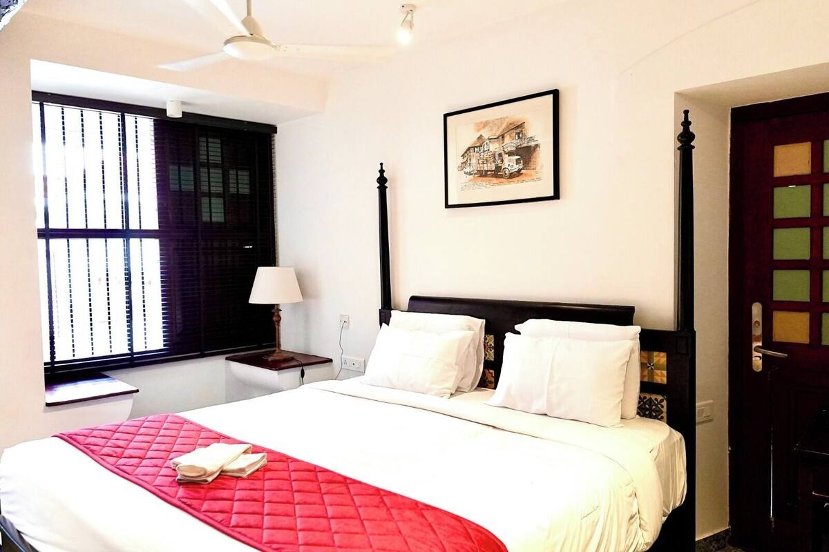 Una habitación de hotel con cama, ventana con persianas, mesita de noche con lámpara y puerta con un panel decorativo.