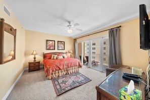 Une chambre à coucher comprenant un lit, des lampes de chevet, un ventilateur de plafond, une grande fenêtre avec des rideaux, un balcon et un téléviseur.