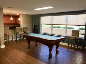 A pool table in a room with a view through a large window, bar stools, and a kitchen area.