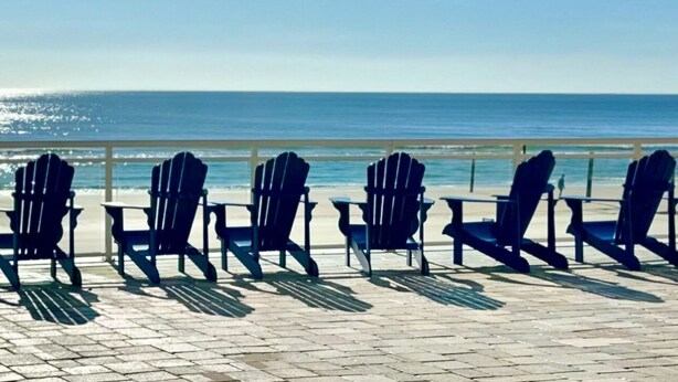 A beachside view with four Adirondack chairs aligned on a paved walkway, overlooking the ocean.