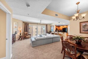 A spacious living area with a large sofa, dining table, and chairs, a kitchen in the background, and a balcony with curtains.