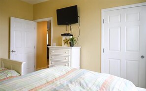 Une chambre à coucher avec un lit, une commode, un téléviseur fixé au mur et deux portes.