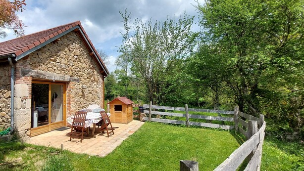 Ein Haus aus Stein mit einer gefliesten Terrasse, Holzmöbeln und einem hölzernen Spielhäuschen.