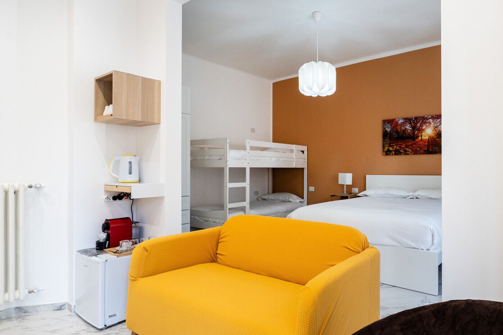 Une chambre d’hôtel compacte comprenant un lit superposé, un lit simple, une petite kitchenette, un fauteuil jaune et un tableau accroché au mur.