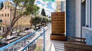 Un balcón con un banco de madera, barandilla metálica y vista a una calle con coches y árboles.
