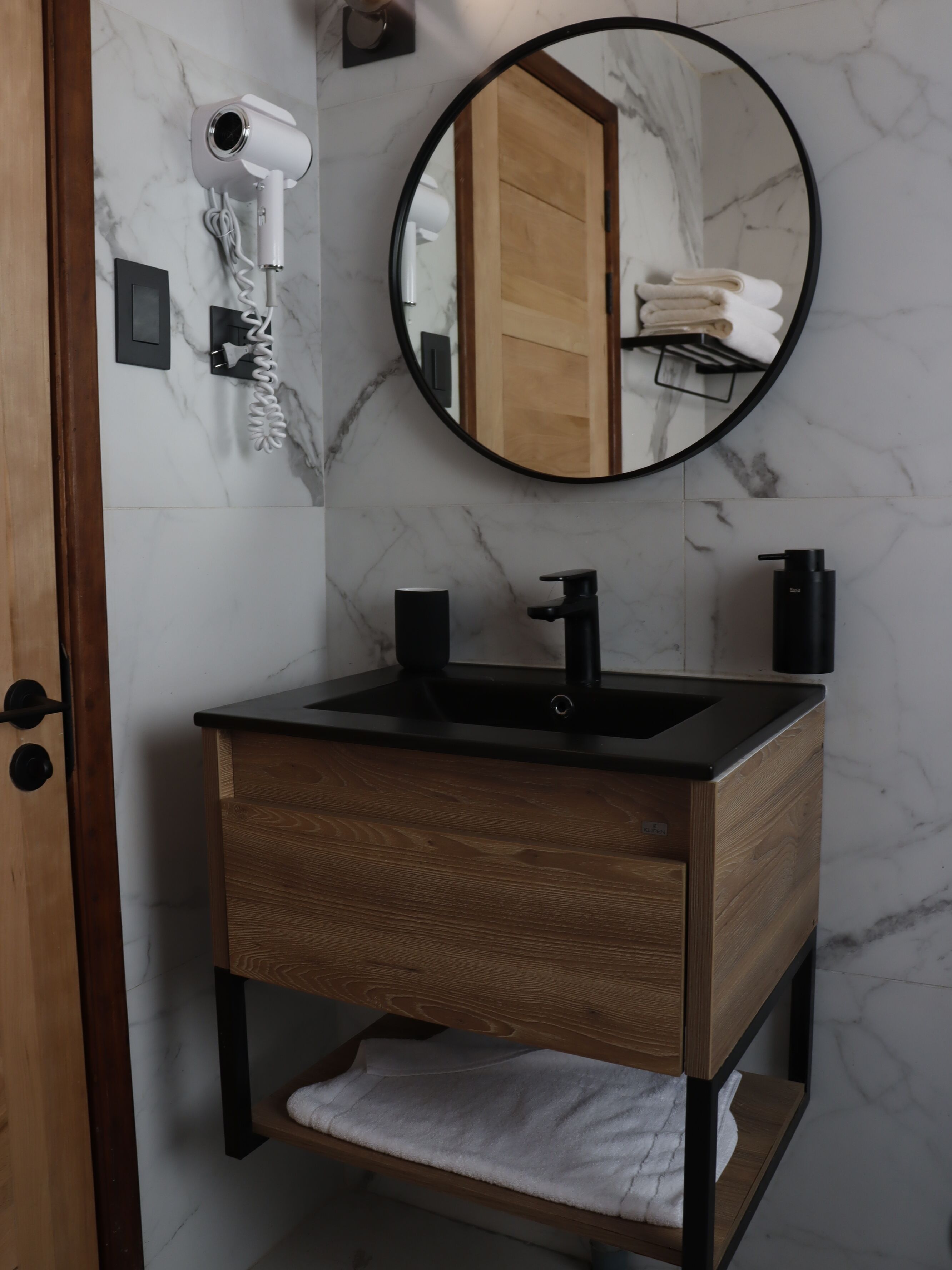Een badkamer met een houten wastafel, een zwarte wasbak, een spiegel en een aan de muur bevestigde haardroger.
