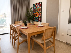 A dining area with a wooden table, six chairs, and a colorful abstract painting on the wall. - Solara - Stylish Balcony Apartments in Larnaca (Larnaca)