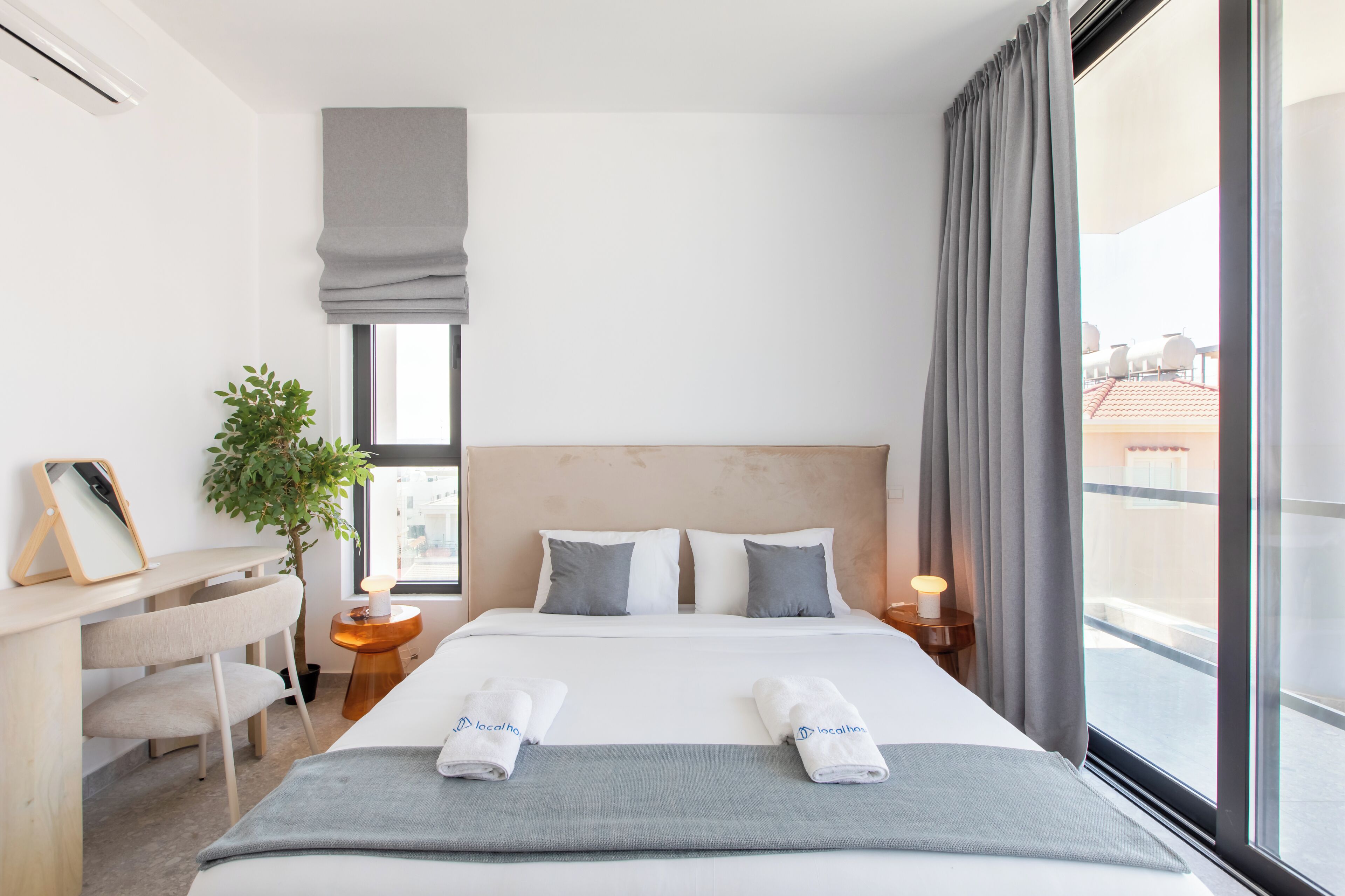 A hotel room with a bed, bedside table, lamp, and a view of the outside.
