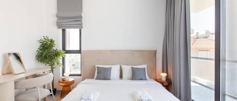 A hotel room with a bed, bedside table, lamp, and a view of the outside.