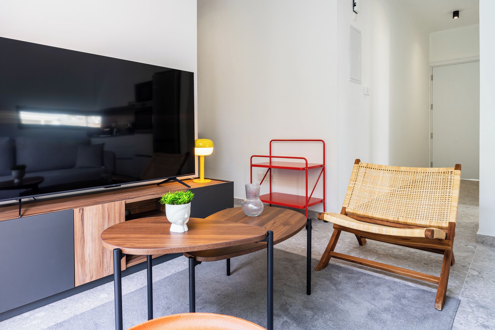 Un salon moderne avec un téléviseur à écran plat, une table en bois, un fauteuil en osier, une étagère en métal rouge et une petite plante en pot.