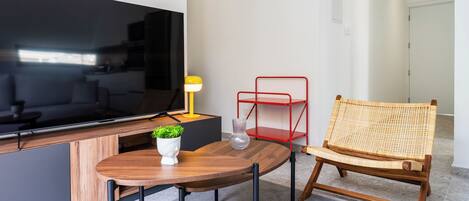 Un salon moderne avec un téléviseur à écran plat, une table en bois, un fauteuil en osier, une étagère en métal rouge et une petite plante en pot.