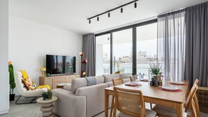 A modern living room with a sofa, dining table, and chairs. A flat-screen TV is mounted on the wall, and there is a view of the city through large windows. - Solara - Stylish Balcony Apartments in Larnaca (Larnaca)