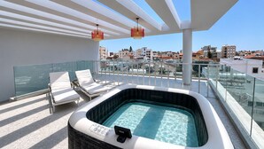 A rooftop terrace with a hot tub, lounge chairs, and a glass balustrade overlooking a cityscape. - Solara - Stylish Balcony Apartments in Larnaca (Larnaca)