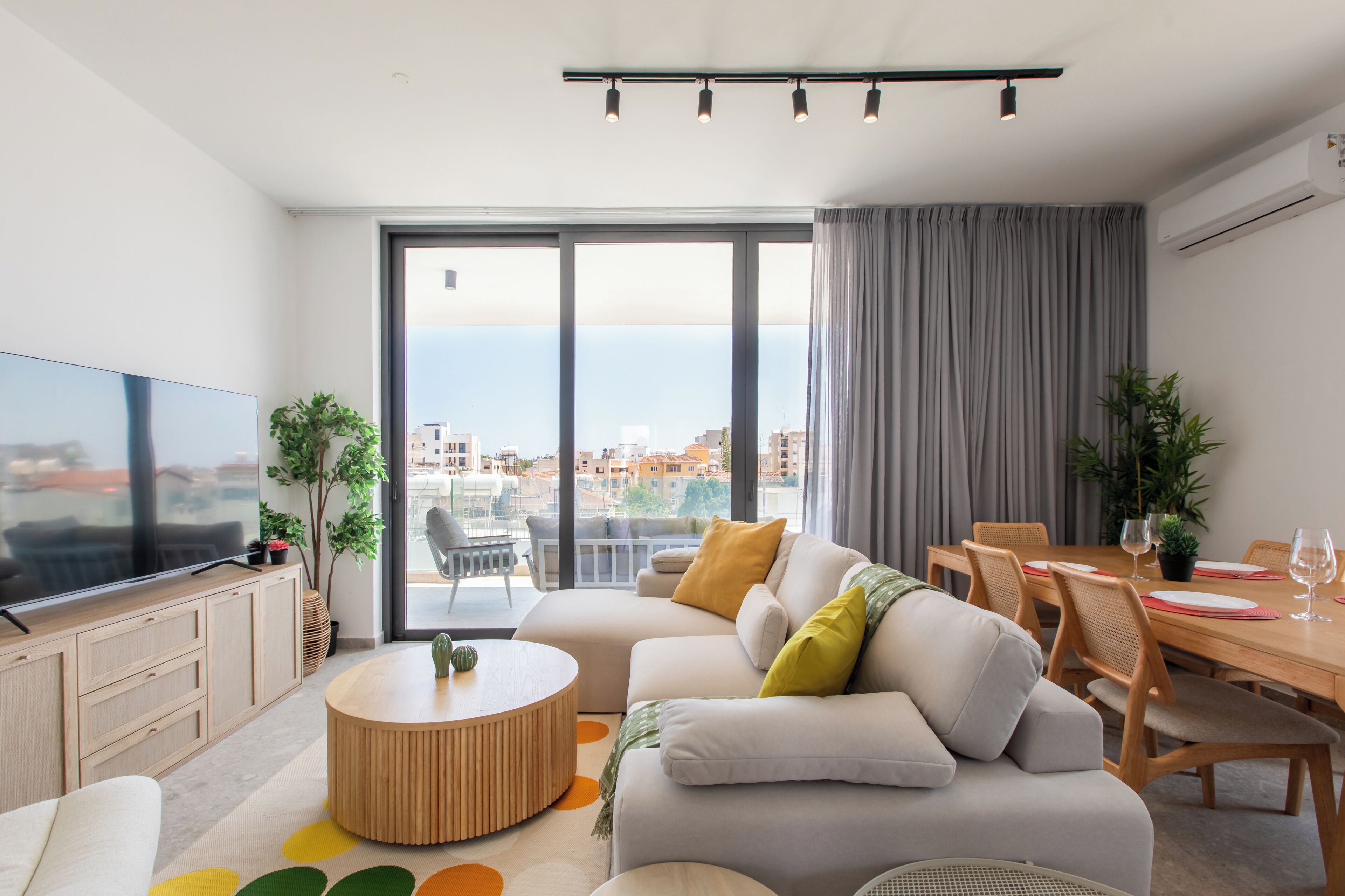 A modern living room with a flat-screen TV, a round coffee table, a sofa, a dining table with chairs, and a view of the city through large windows.