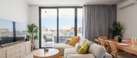 A modern living room with a flat-screen TV, a round coffee table, a sofa, a dining table with chairs, and a view of the city through large windows.
