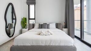 A modern hotel room with a large bed, a mirror, a potted plant, and a view of the city. - Solara - Stylish Balcony Apartments in Larnaca (Larnaca)