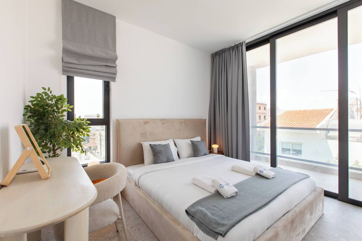 A modern hotel room with a large bed, a desk, a potted plant, and a view of the city.