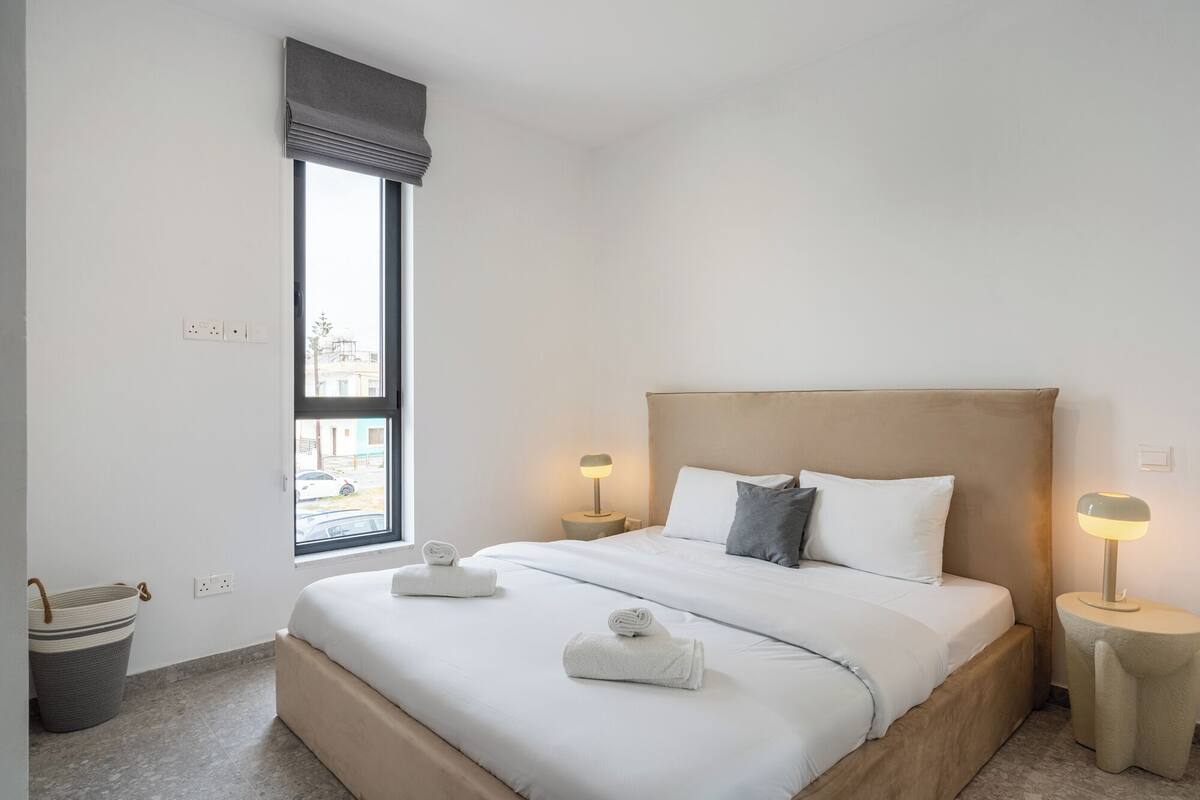 A hotel room with a large bed, two bedside lamps, a window with a view, and a small trash bin.