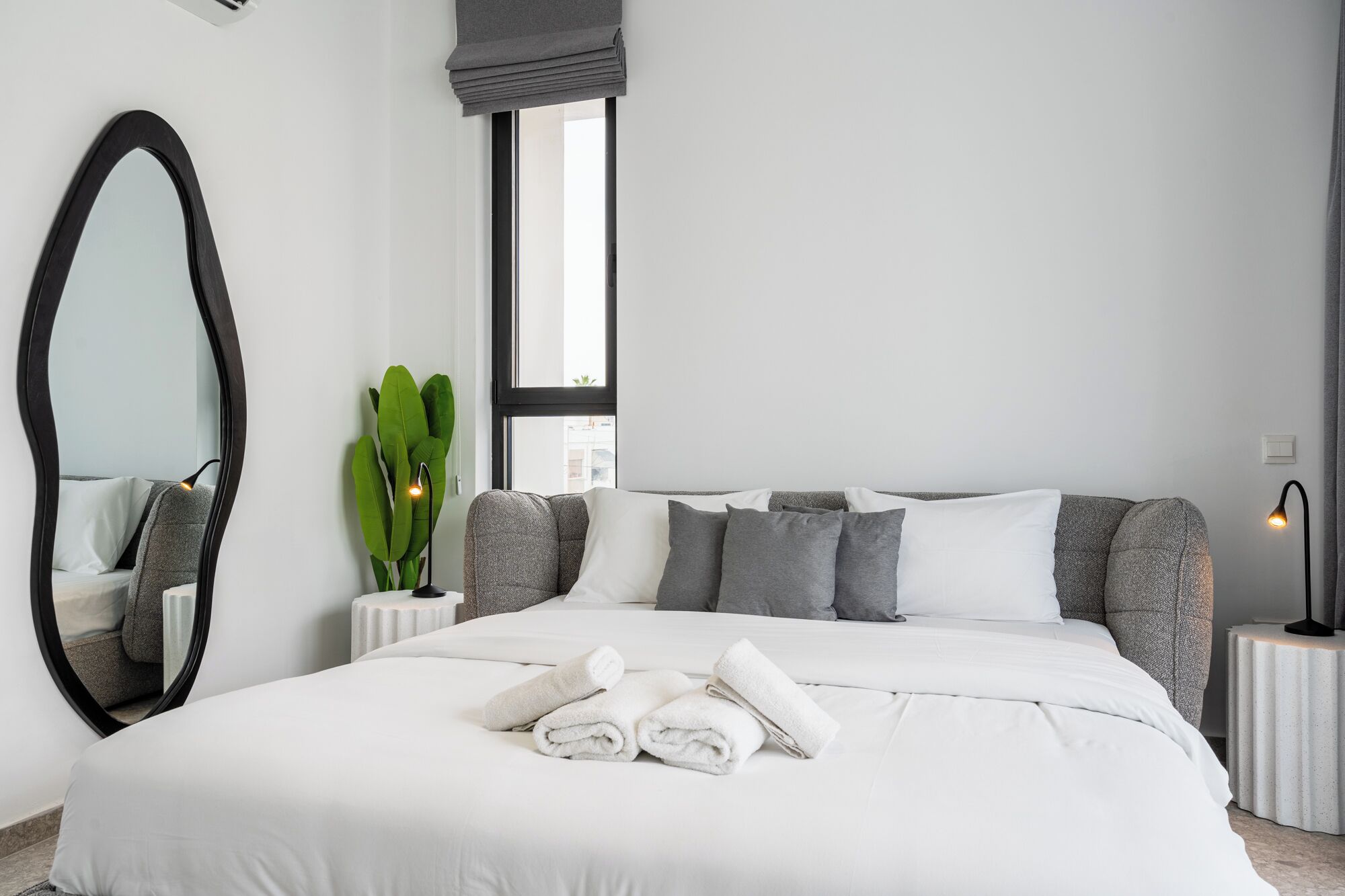 A modern bedroom with a large bed, a round mirror, a potted plant, and bedside lamps.