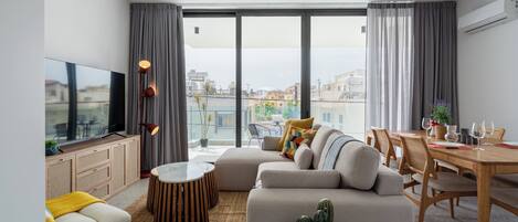 A modern living room with a sofa, a coffee table, a dining table with chairs, a TV, and a large window with curtains.