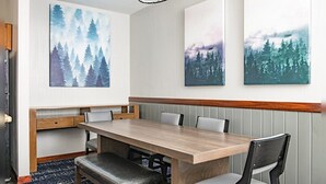 Zona pranzo con tavolo in legno, panca imbottita e sedie. Tre grandi quadri a tema forestale adornano la parete.