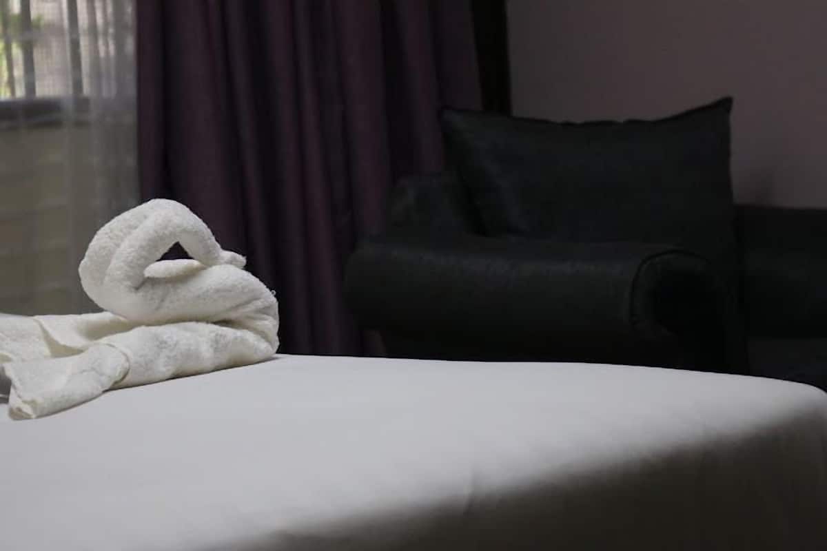 A neatly made bed with a white towel on the left side, a dark-colored sofa, and a window with curtains in the background.