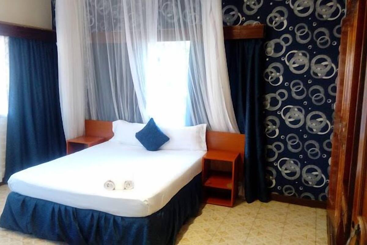 A hotel room with a bed, a nightstand, a window with sheer curtains, and a patterned wall.