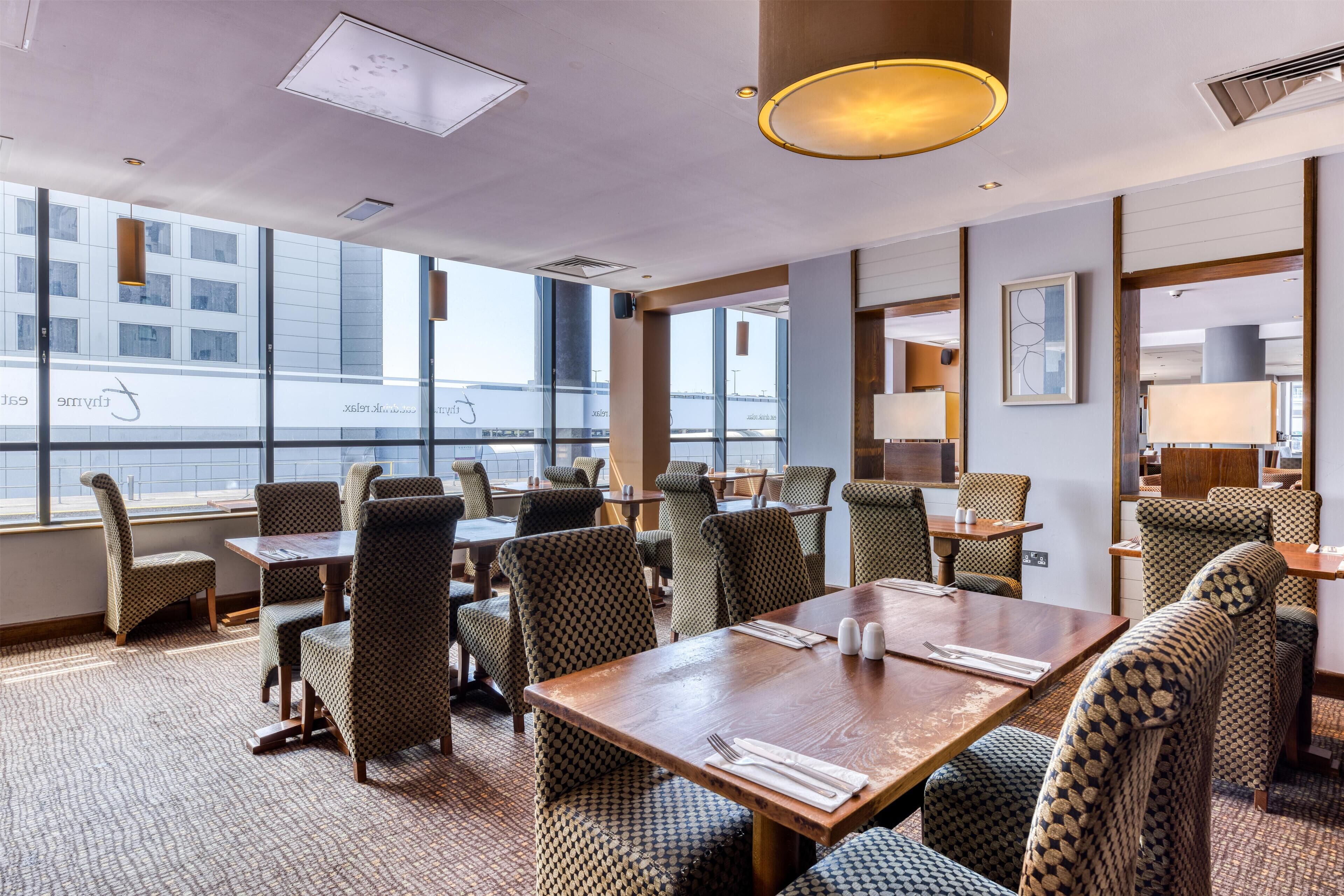 A hotel dining area with patterned chairs, wooden tables, and large windows offering a view of a building.