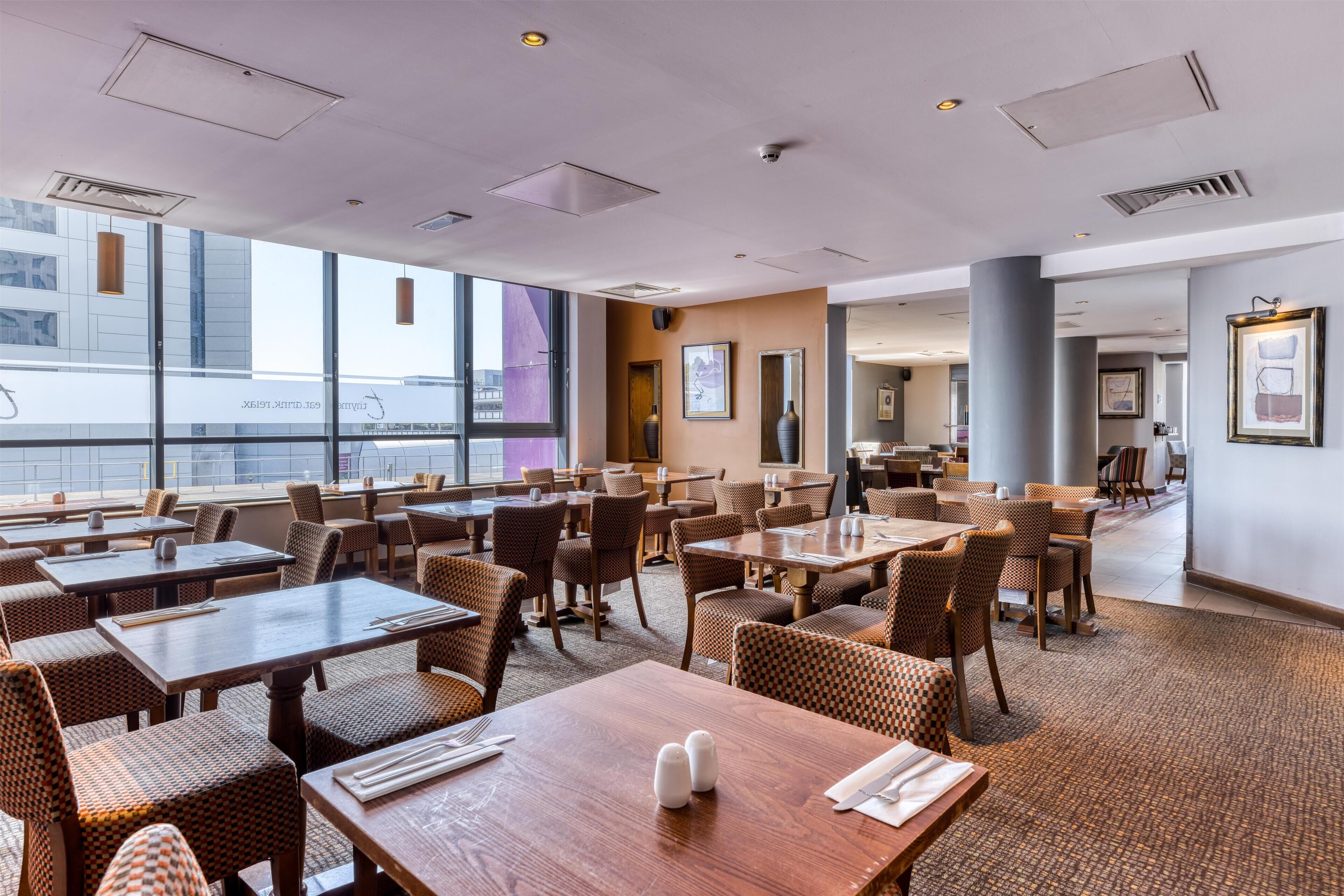 A modern restaurant with high ceilings, large windows, and a mix of wooden and wicker furniture.