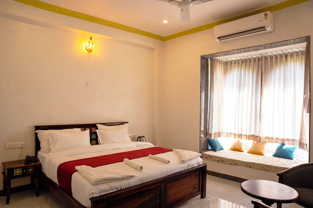 A hotel room with a bed, bedside table, air conditioning unit, window with curtains, and a small table.