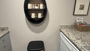 Una cocina con un cubo de basura negro, una estantería de pared con tazas y un cuadro en la pared.