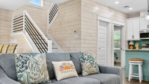 A modern living room with a grey sofa, decorative pillows, a staircase, and a kitchen area. - Coastal Retreat (Rockport)