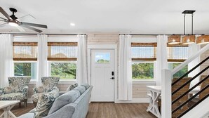 A spacious living room with a grey sofa, patterned armchairs, a wooden floor, a white staircase, and large windows with white curtains. - Coastal Retreat (Rockport)