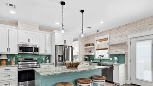 A modern kitchen with white cabinets, stainless steel appliances, teal countertops, and three bar stools. - Coastal Retreat (Rockport)