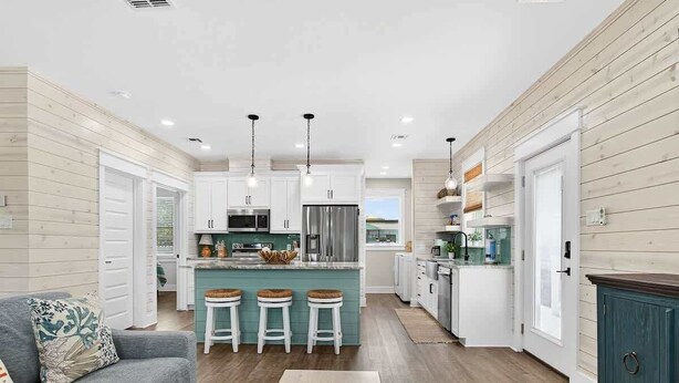 A modern kitchen with white cabinets, a central island with stools, and a grey sofa in a living area. - Coastal Retreat (Rockport)