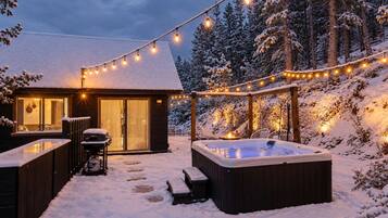 A snow-covered outdoor area with a hot tub, string lights, and a wooden cabin.