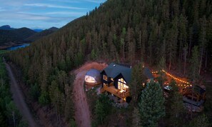 A cabin with a large dome structure, surrounded by dense forest and a dirt road. - Geo Dome Group Retreat | 3 King Beds & Game Room (Bailey)