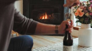 A person sitting by a fireplace with a bottle and flowers on the table.