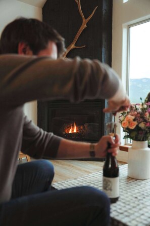 A person sitting by a fireplace with a bottle and flowers on the table. - Geo Dome Group Retreat | 3 King Beds & Game Room (Bailey)