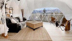 A cozy interior with a sofa, ottoman, and chairs, a large window with a mountain view, and a hanging light fixture.