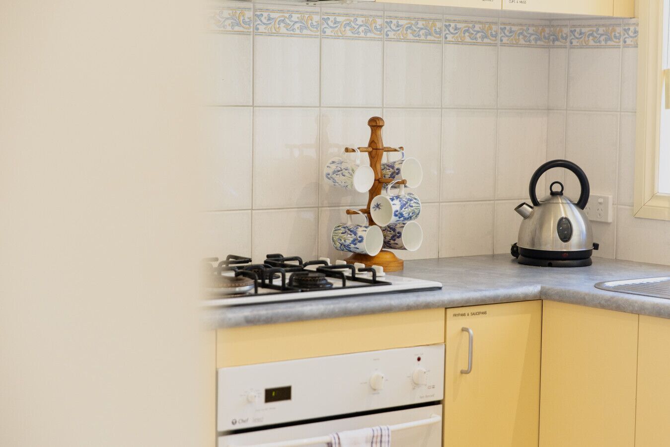 Una cocina con estufa a gas, hervidor y un juego de té decorativo sobre una estantería.