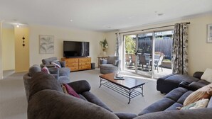 Uma sala de estar moderna com uma televisão de tela plana grande, sofás cinza, uma mesa de centro de madeira e uma porta de vidro deslizante que dá para um pátio externo.