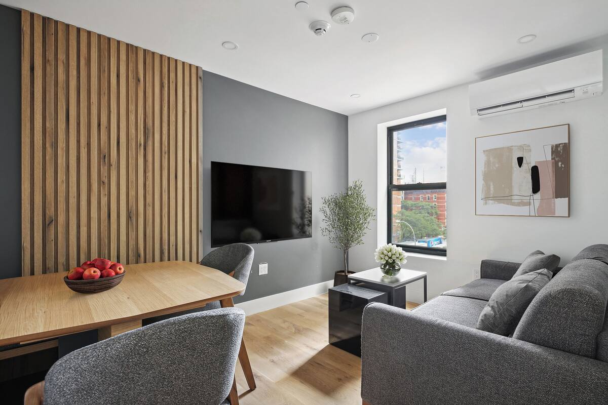 A modern living room with a wooden panel wall, a flat-screen TV, a dining table with a bowl of fruit, grey upholstered furniture, and a window with a view of buildings.