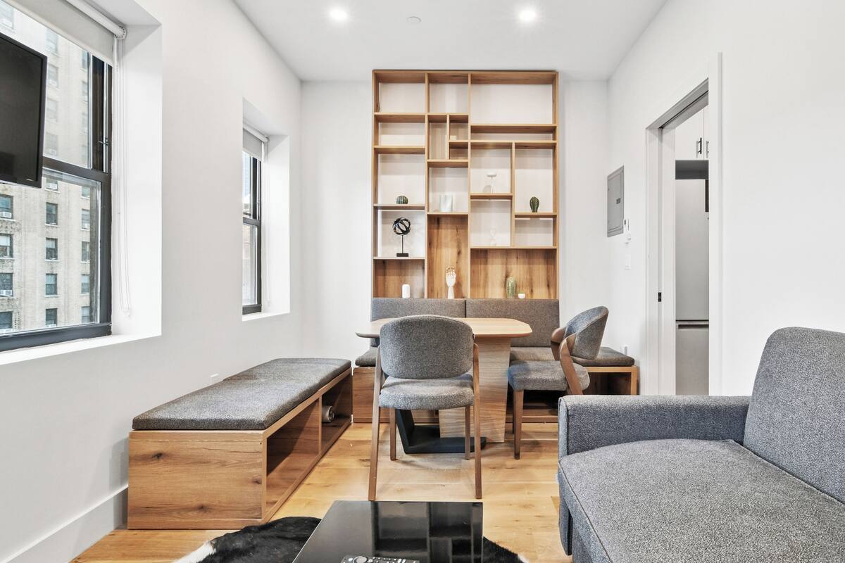 A modern living room with a wooden bench, grey upholstered chairs, a black coffee table, and a built-in wooden shelving unit.