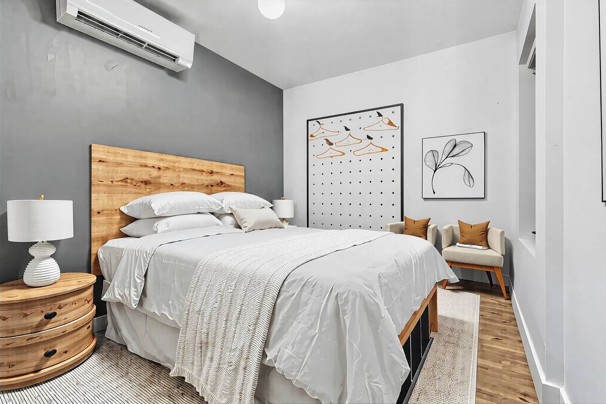A bedroom with a wooden headboard, a bed with white bedding, a wall-mounted air conditioner, a wall art piece, and a side table with a lamp.